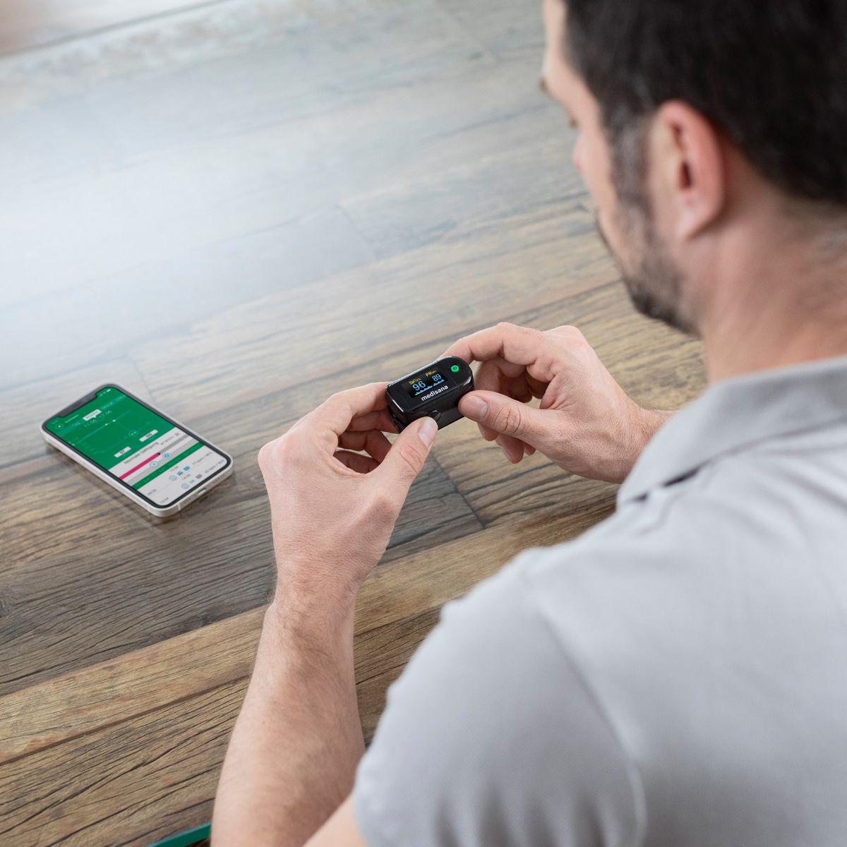Ein Mann hält ein schwarzes Medisana Pulsoximeter in den Händen, während neben ihm ein Smartphone mit der passenden Gesundheits-App liegt.